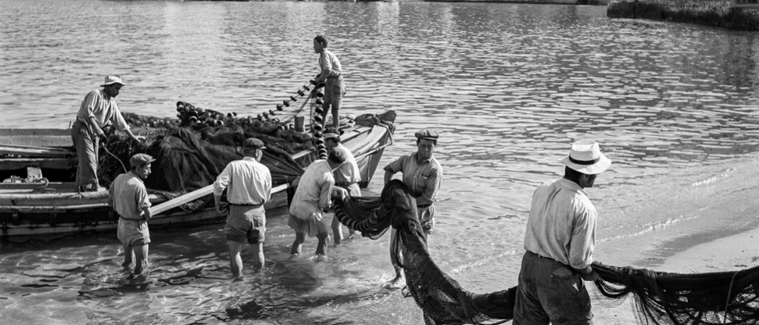 Μύκονος, 1955. Ξημερώματα στον μόλο Καμπάνη, ψαράδες ξεφορτώνουν τα δίχτυα τους για στέγνωμα και επισκευή. Το νεόκτιστο ξενοδοχείο Λητώ διαφαίνεται στο βάθοςi