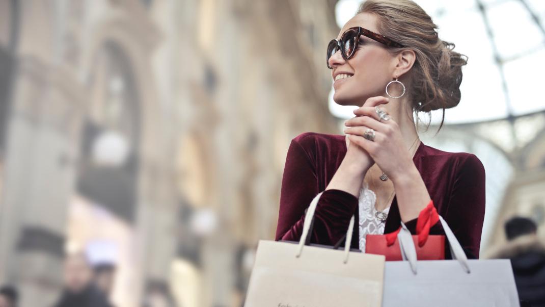 Γυναίκα με γυαλιά ηλίου χαμογελάει και κρατάει σακούλες με ψώνια