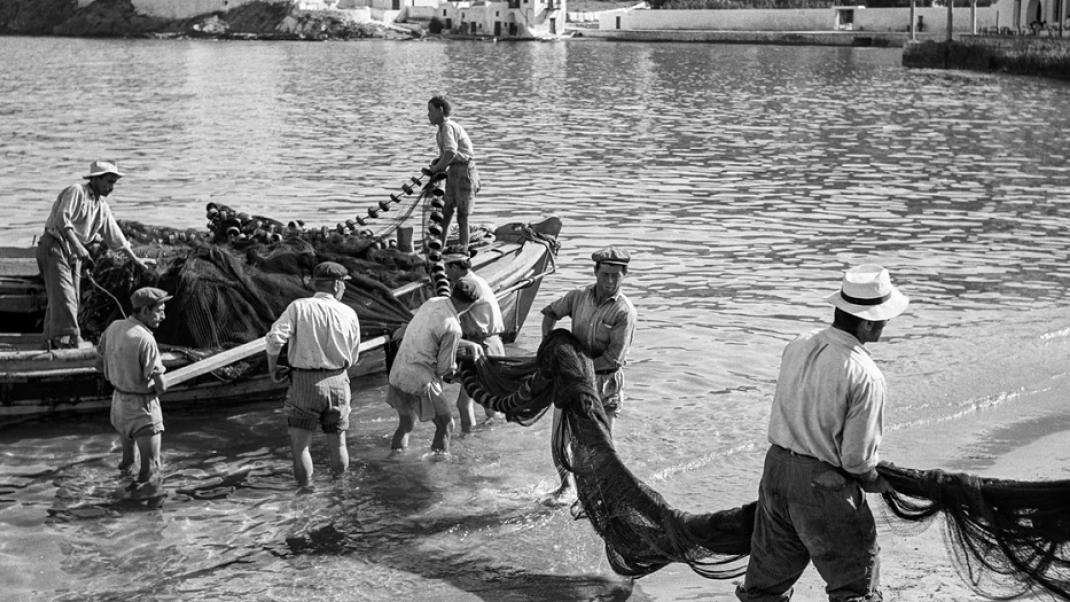 Μύκονος, 1955. Ξημερώματα στον μόλο Καμπάνη, ψαράδες ξεφορτώνουν τα δίχτυα τους για στέγνωμα και επισκευή. Το νεόκτιστο ξενοδοχείο Λητώ διαφαίνεται στο βάθοςi