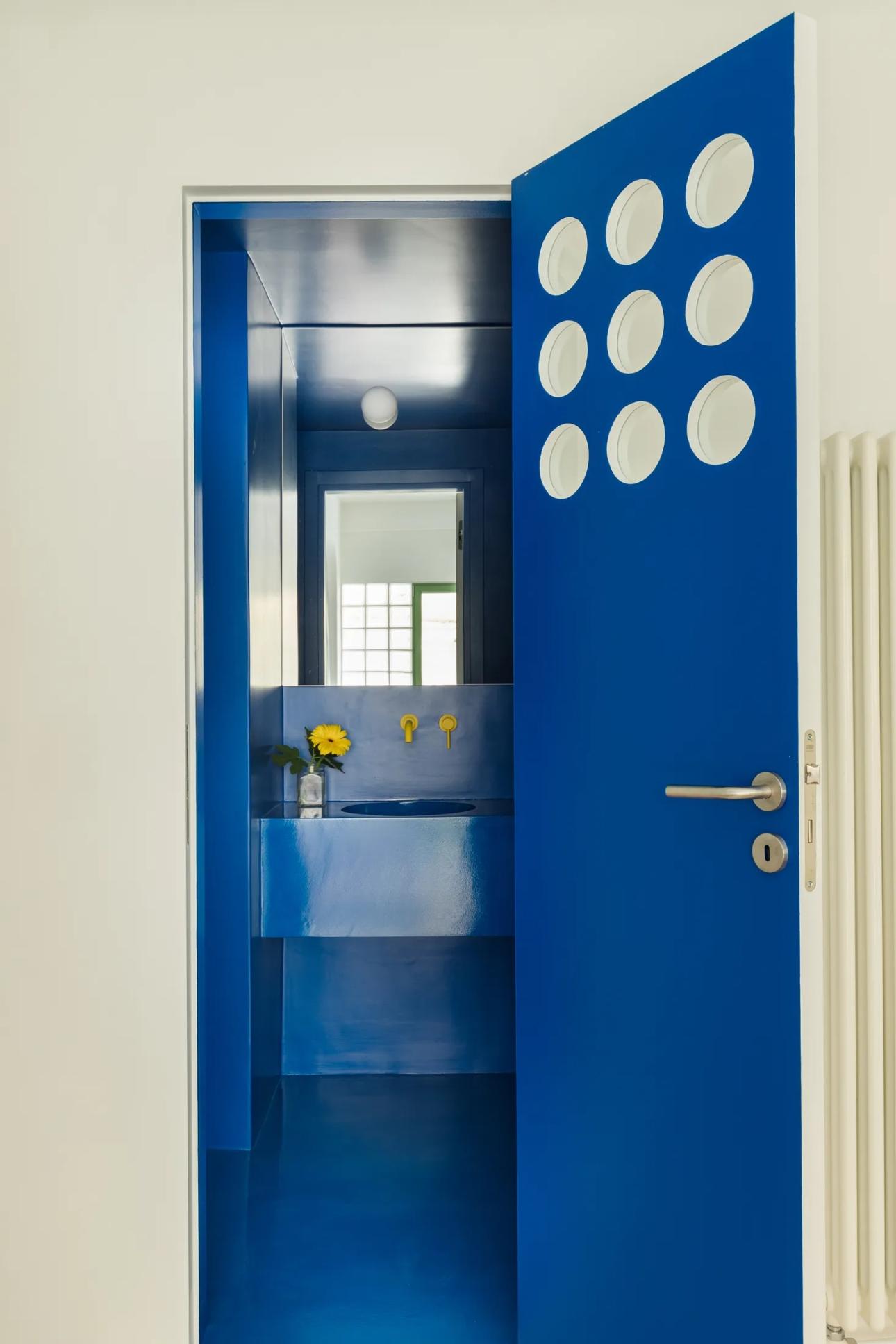 Το μπάνιο φωτίζεται έμμεσα μέσα από την πόρτα με τα κυκλικά ανοίγματα, μετατρέποντας την έλλειψη παραθύρου σε σχεδιαστικό πλεονέκτημα.