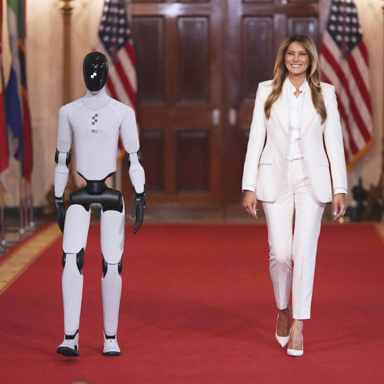 Η Μελάνια Τραμπ με λευκό κοστούμι περπατάει στον λευκό οίκο δίπλα σε ένα ρομπότ