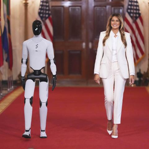Η Μελάνια Τραμπ με λευκό κοστούμι περπατάει στον λευκό οίκο δίπλα σε ένα ρομπότ